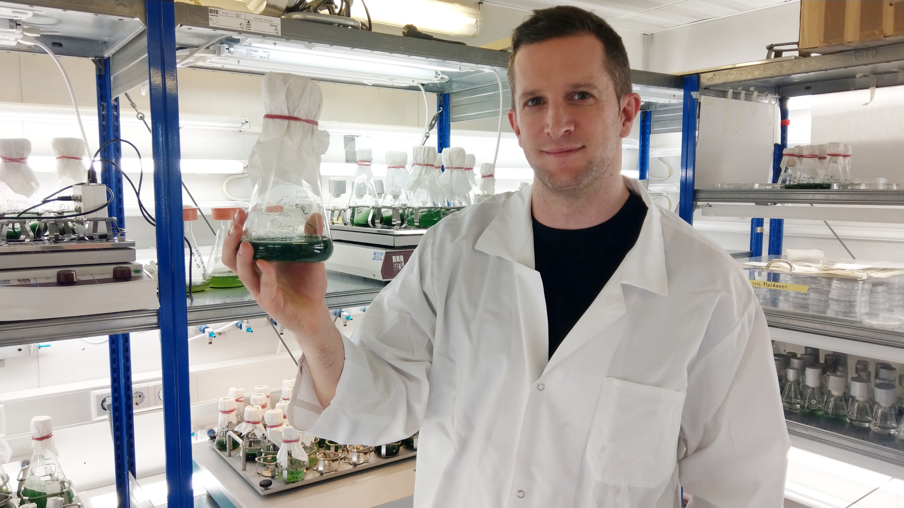 The photo shows the researcher in his laboratory holding an Erlenmeyer flask containing a green cyanobacteria culture