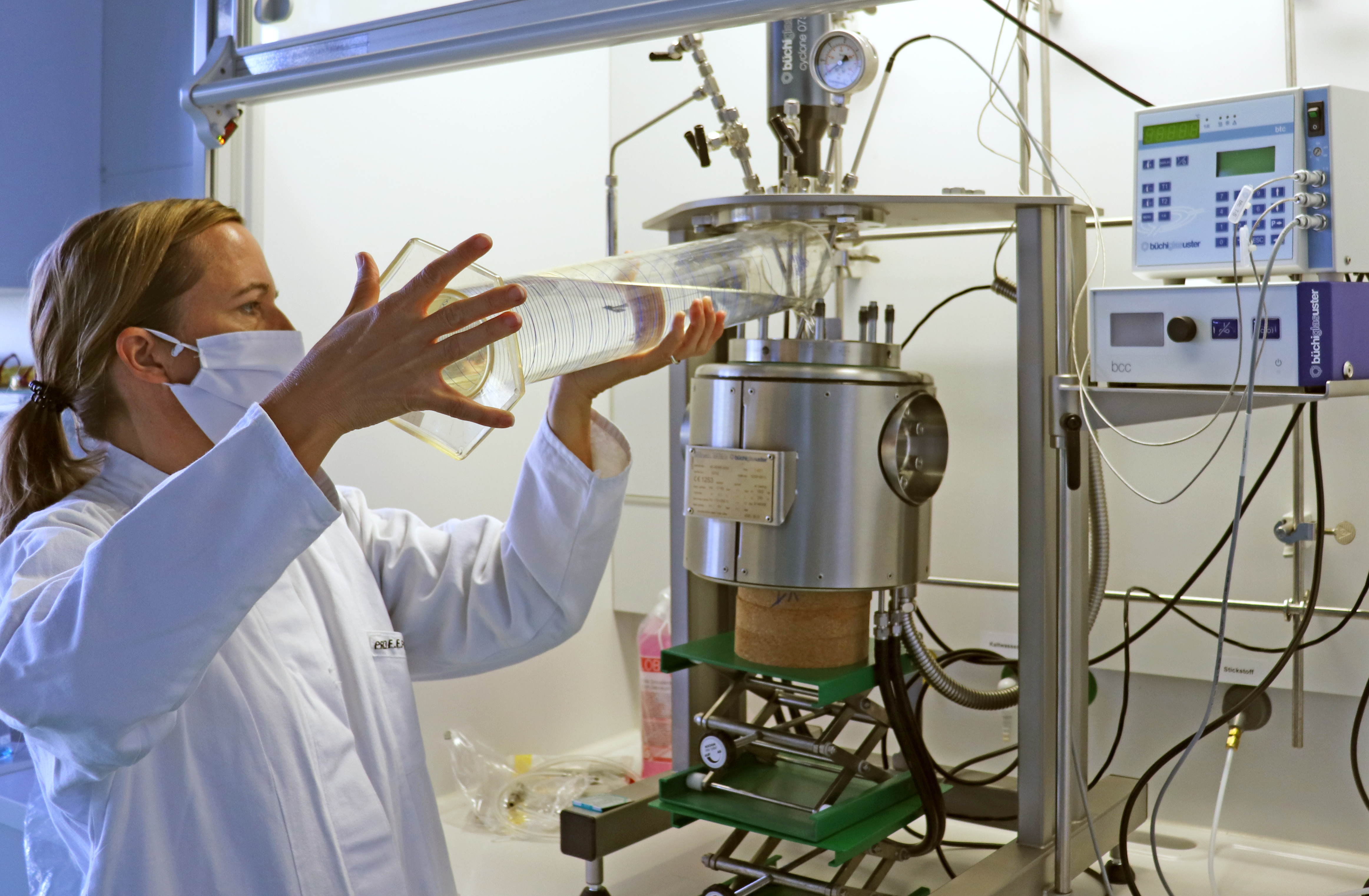Zu sehen ist eine Frau in einem weißen Laborkittel vor einer Apparatur aus Edelstahl. Die Frau füllt aus einem großen Messzylinder eine klare Flüssigkeit in den Edelstahlbehälter der Apparatur.