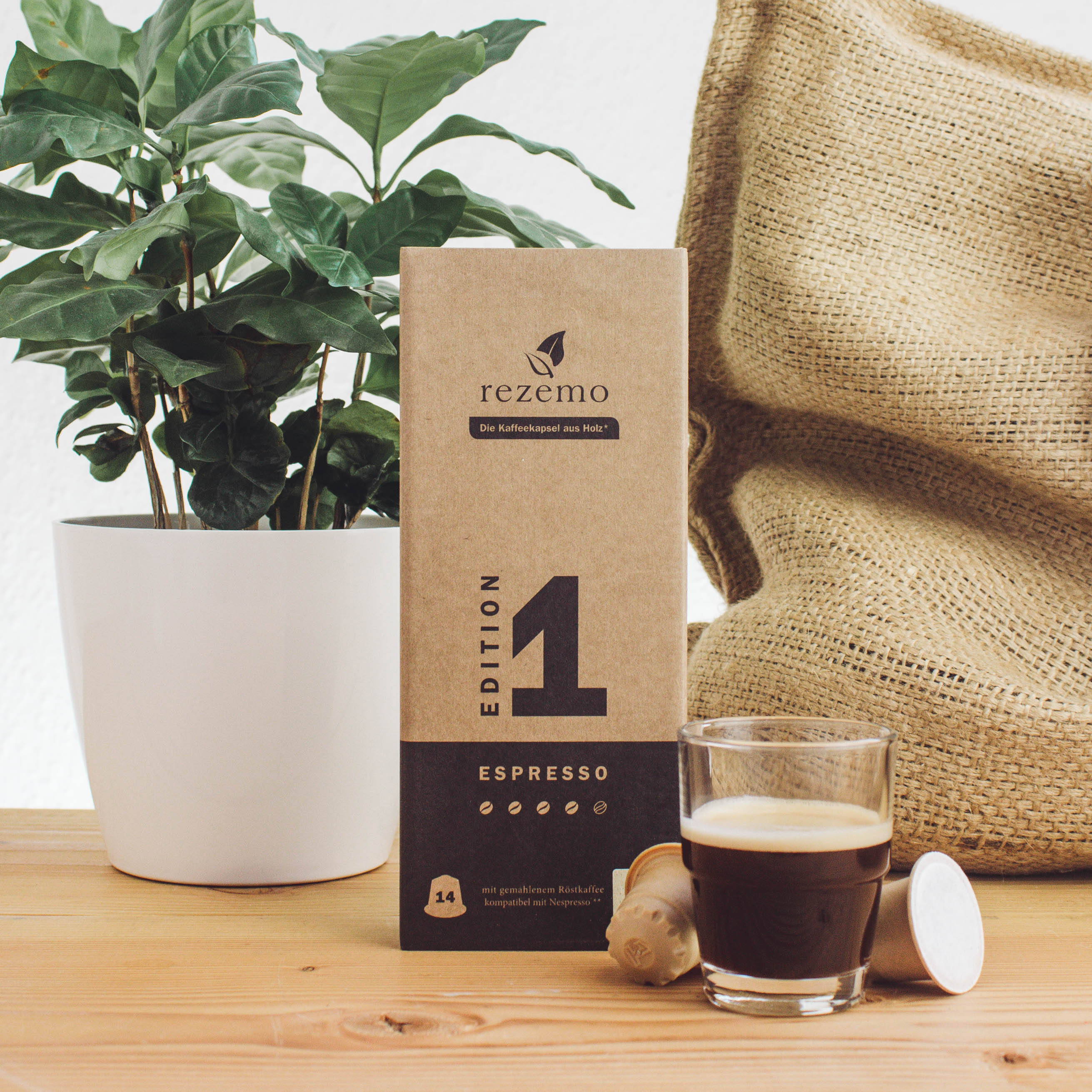 Eine Kaffeepflanze, eine rezemo-Verpackung und eine gefüllte Glas-Kaffeetasse vor einem Jutesack.