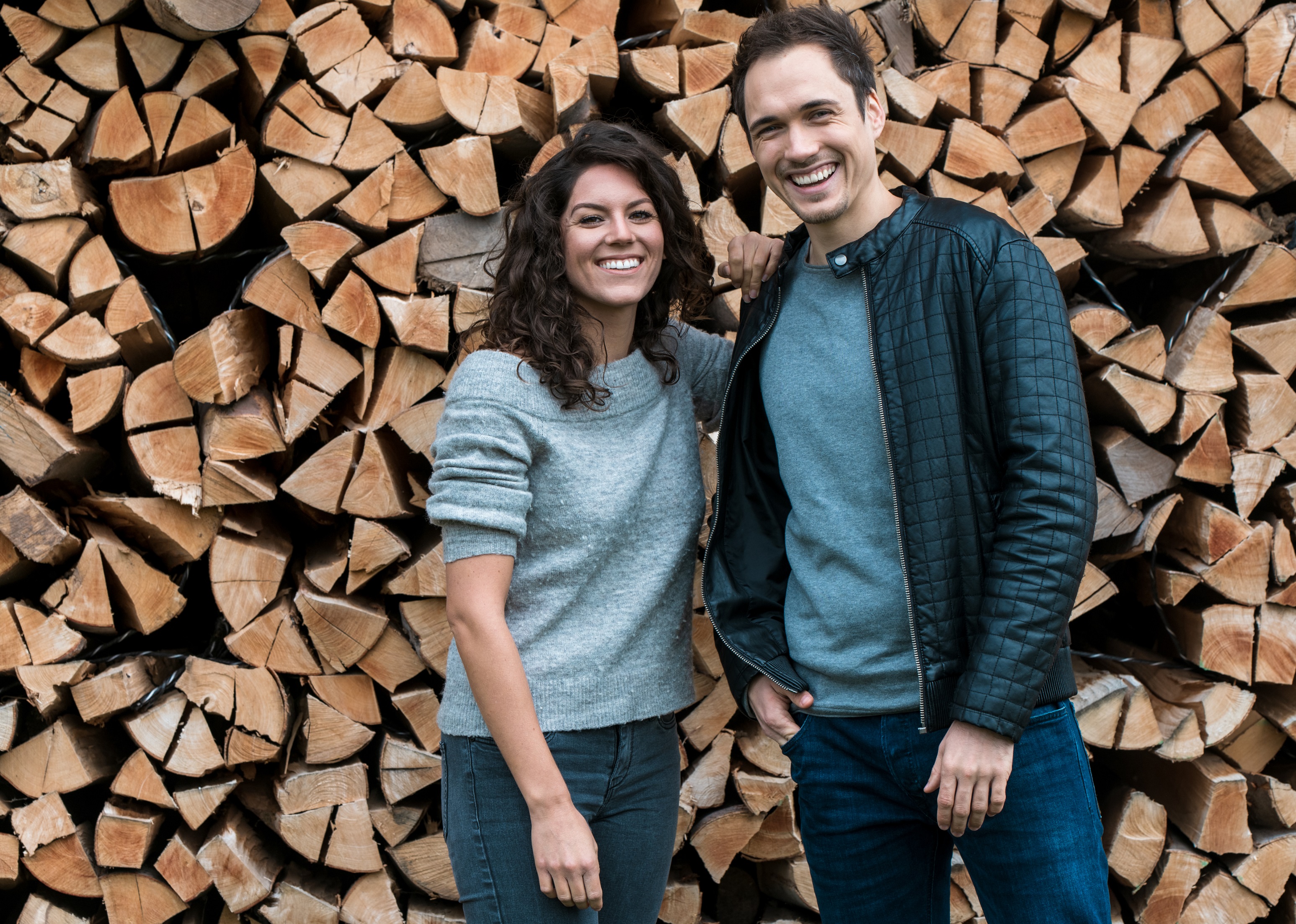 Eine junge Frau und ein junger Mann in der Natur vor gestapeltem Holz.