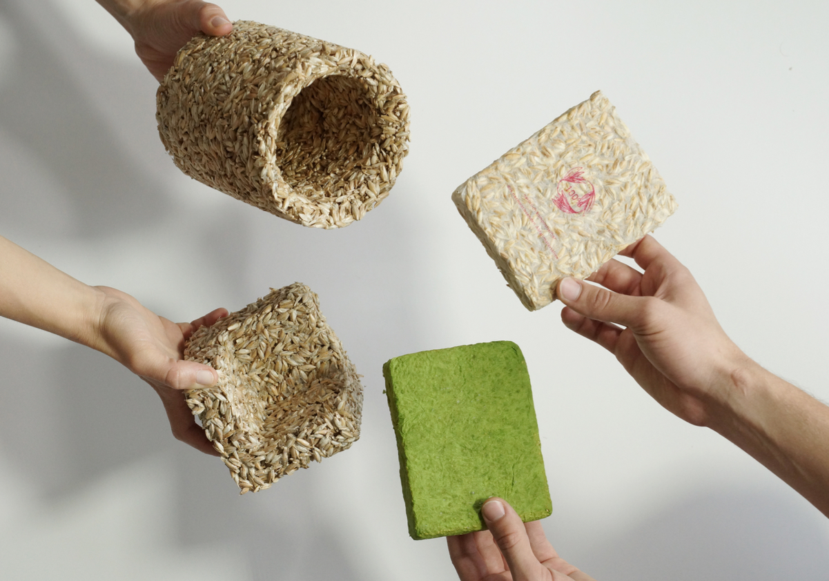 Four hands each hold a different prototype made of husks: a bottle wrapper, an edge protector and a green and light brown block made of the natural material.