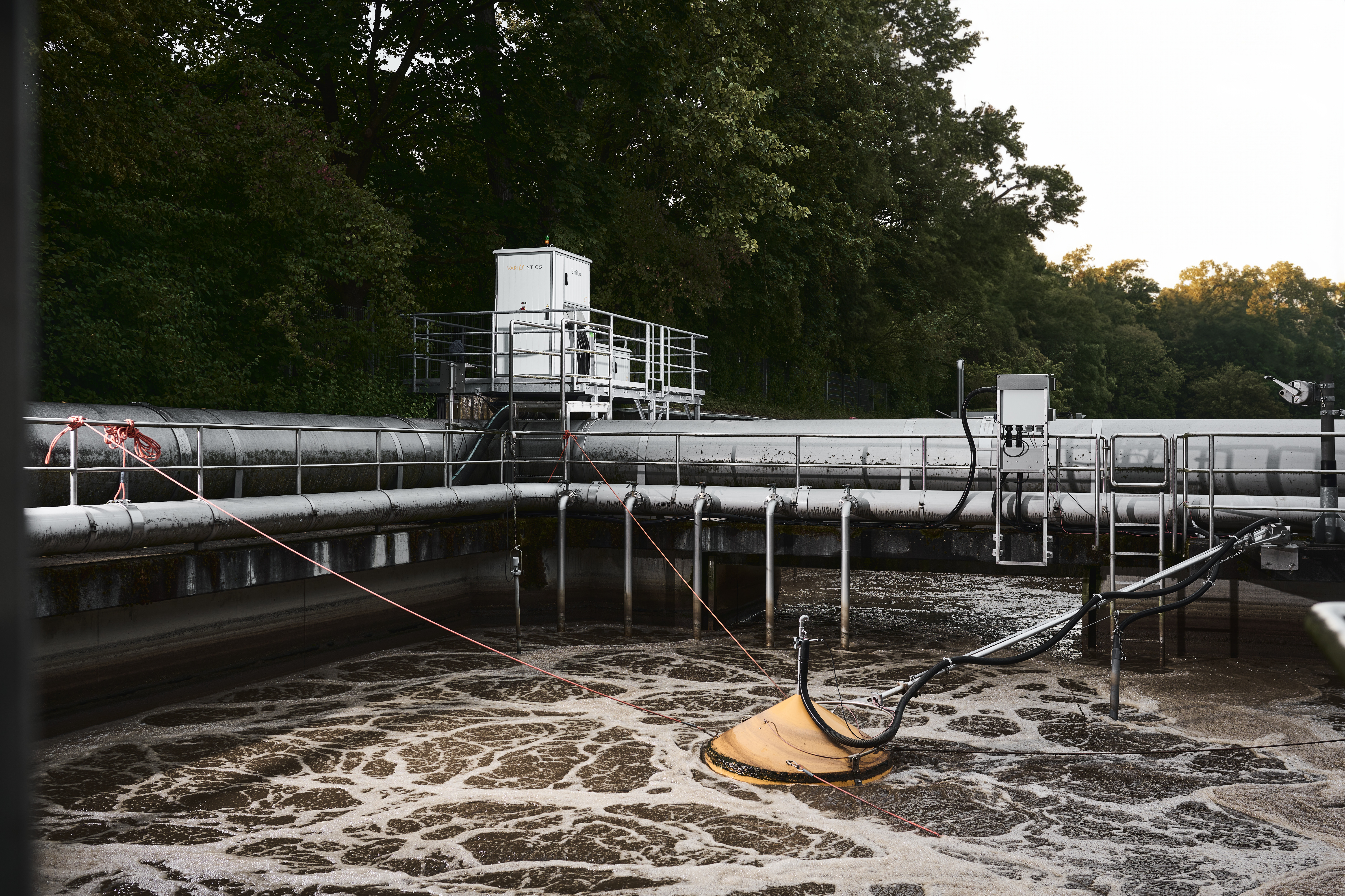 Wastewater treatment plant with a tank of moving wastewater in which an orange hood floats and is connected to an analyser outside the tank.