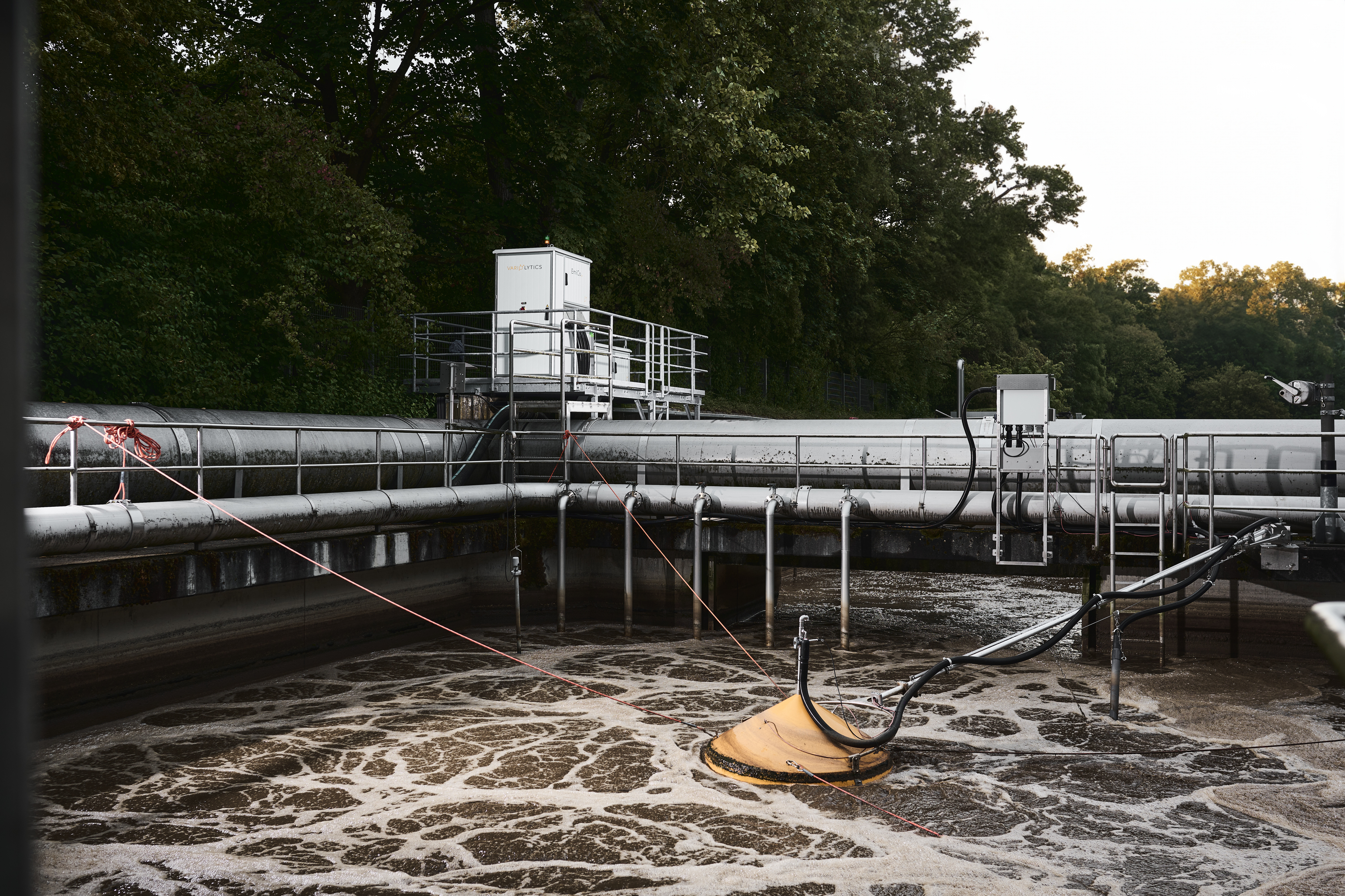 Kläranlage mit einem Becken mit bewegtem Abwasser, in dem ein orangene Haube schwimmt und mit einem Analysegerät außerhalb des beckens verbunden ist.