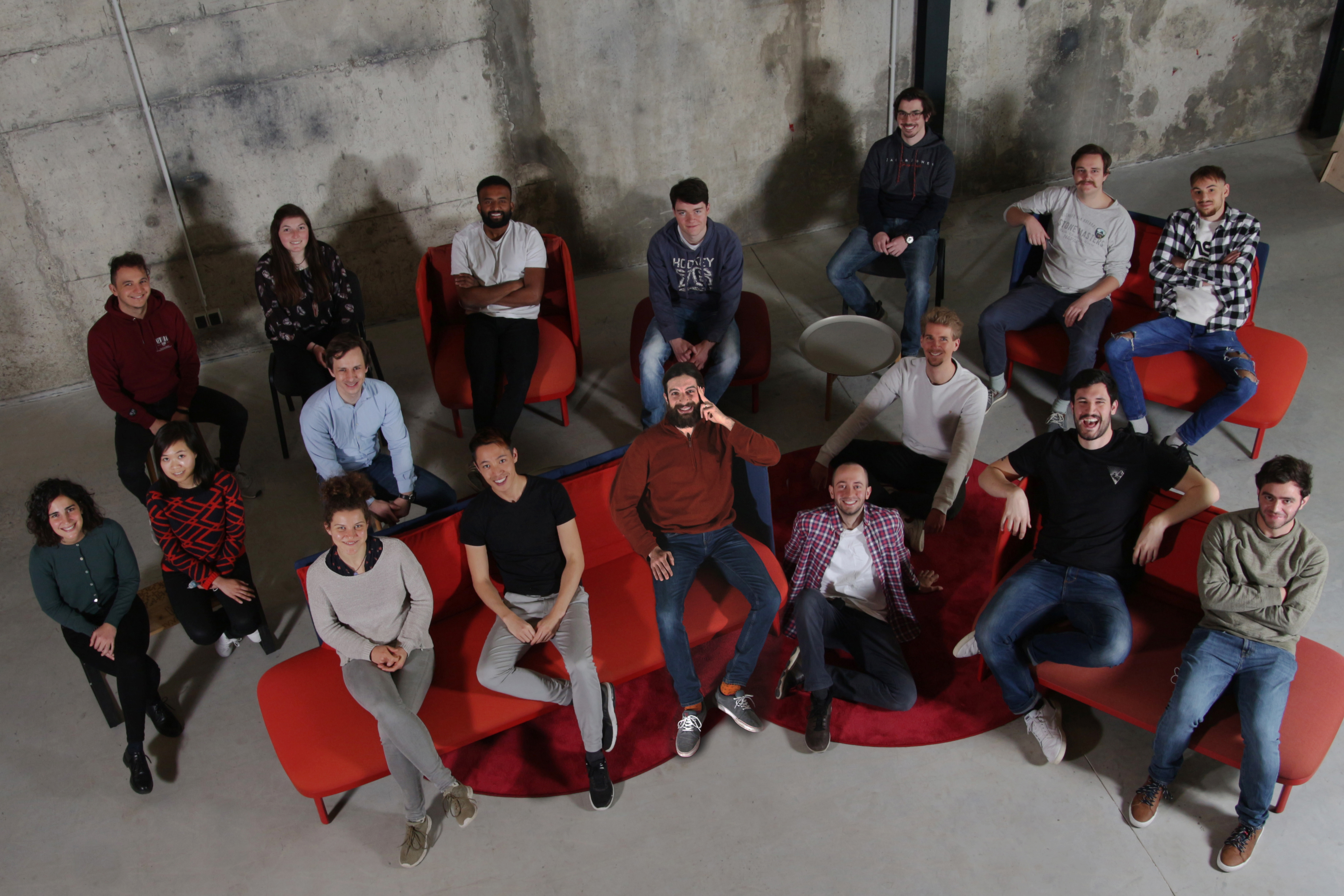 Gruppenbild der Alpha-Protein GmbH: 17 Personen sitzen auf roten, gepolsterten Bänken und Sesseln vor einer Sichtbeton-Wand.