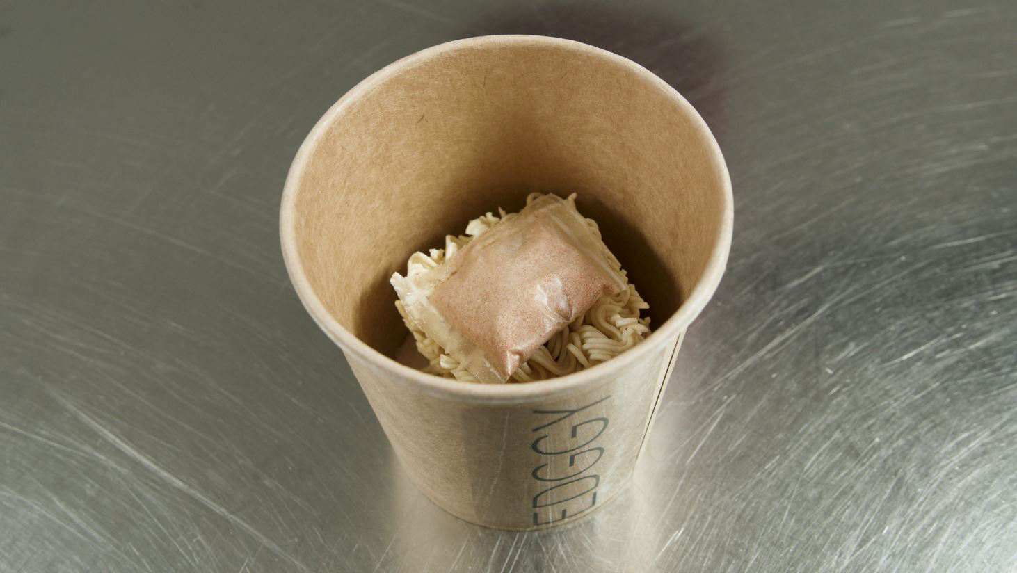 View of a paper cup with dried ramen noodles and a spice sachet on top.