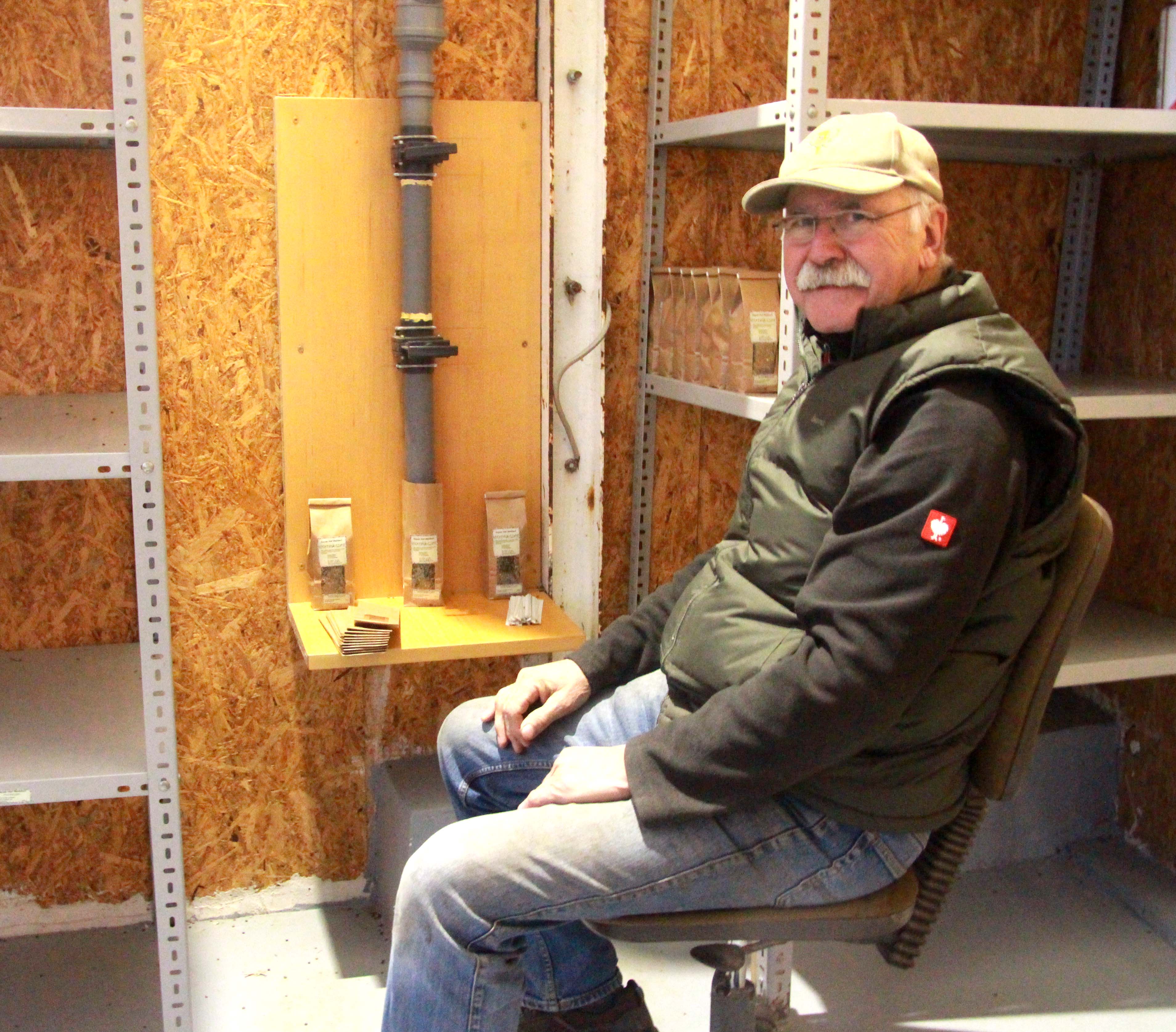 The photo shows a man with a moustache and a cap sitting on a chair at a device with a plastic tube suitable for filling lentils into bags.