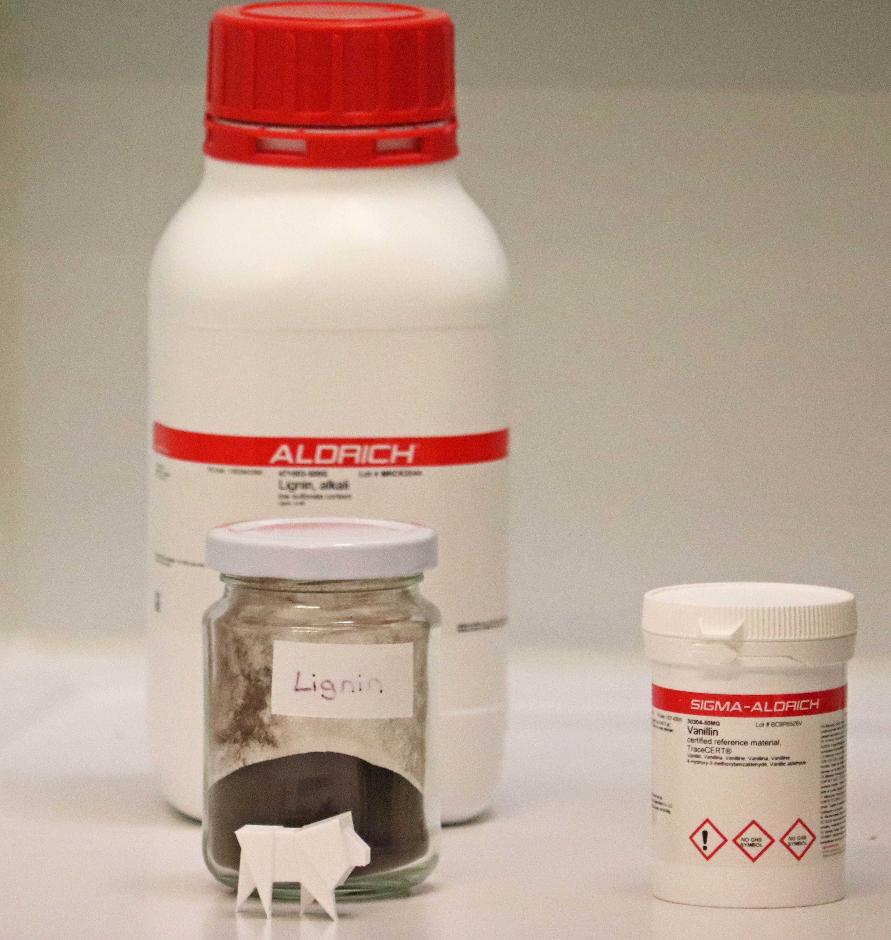 A large white plastic bottle with a red lid and a white-red label, a smaller plastic container with a white lid and a red-white label with black writing, and a glass container with a white lid and a label with the inscription Lignin can be seen. The glass container contains a brown powder.