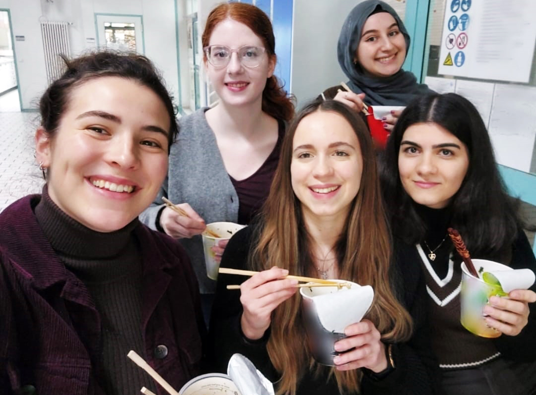 Five young women with instant ramen soups in paper cups and chopsticks in their hands smile friendly into the camera.
