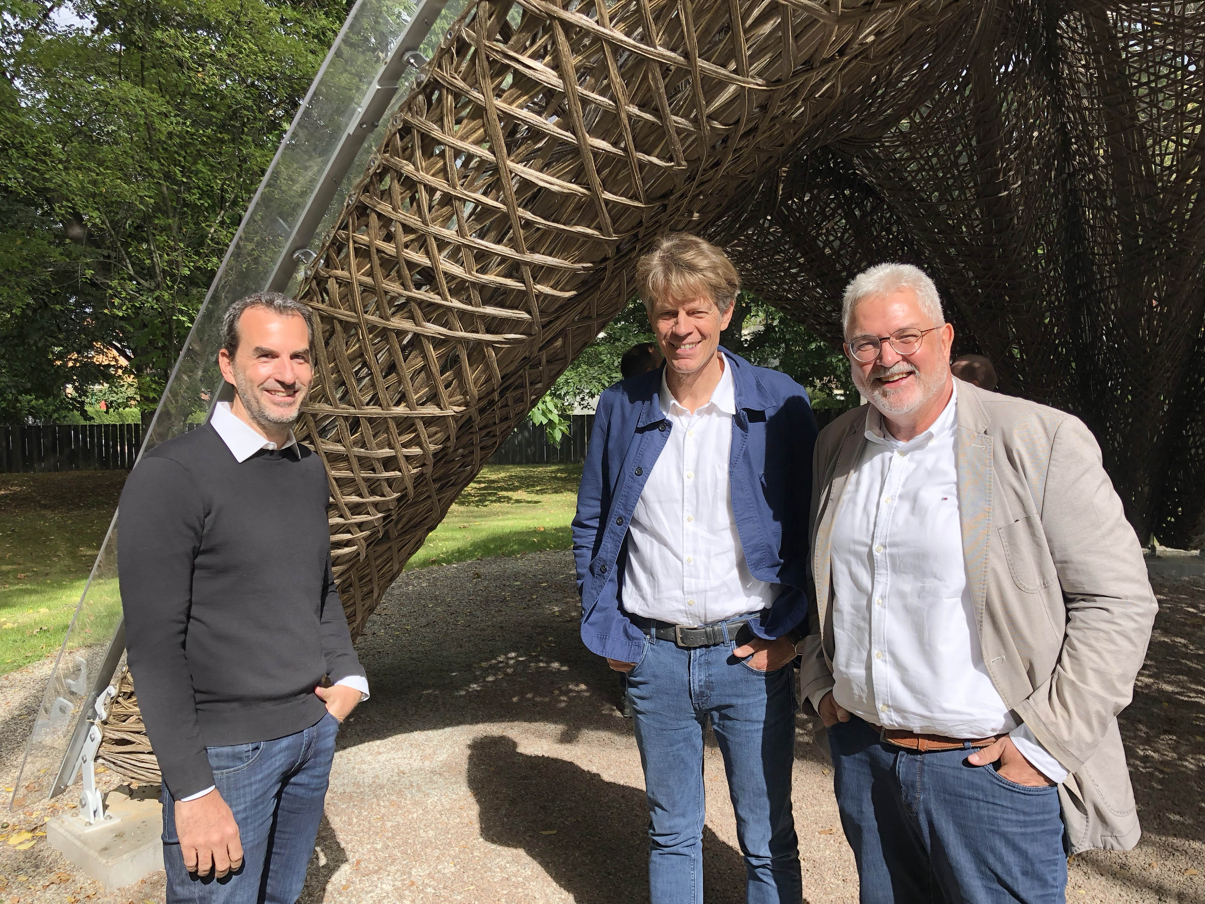 Group picture of three middle-aged men outdoors in front of a support element of the livMatS pavilion