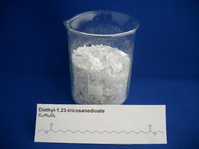 The photo shows a white substance in a beaker. A piece of paper showing the name and the chemical structure of the substance is placed in front of the beaker.