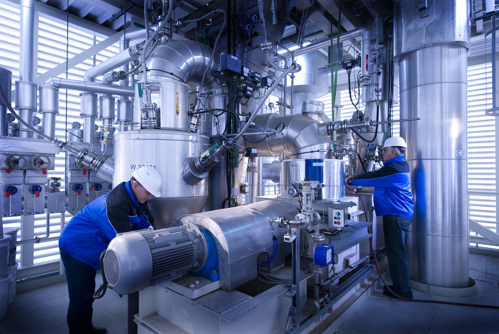 Das Bild zeigt zwei Männer in blauen Jacken, die an einem Reaktor arbeiten.