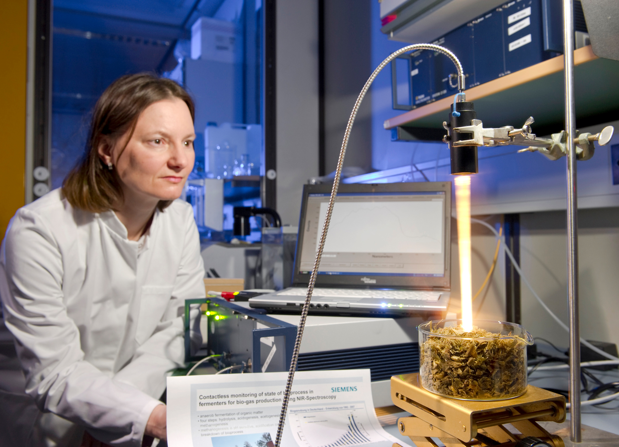 Auf der linken Seite sieht man eine Frau im Laborkittel in einem Labor. Rechts steht ein Laptop und davor ein Becherglas mit Biomasse, welches mit Licht angestrahlt wird.