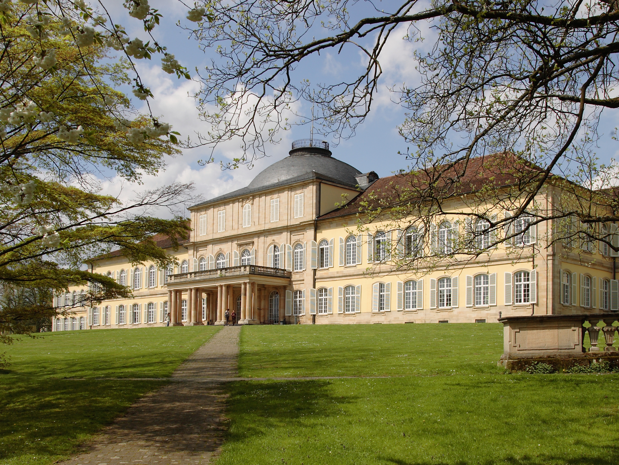 The historic Hohenheim Castle - a sand-coloured pompous building with balcony and windows. It is located in a green area. Blue sky and clouds in the background.