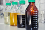 Nebeneinanderstehende Laborflaschen; die Ausgangslösung ist durchsichtig und leicht gelblich, die Kohlenhydratlösung ist gelb-bräunlich, das Prozesswasser ist fast schwarz und das aufgereinigte HMF ist dunkelbraun.