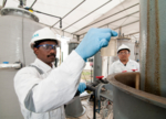 The picture shows two asian employees of the Siemens company in lab coats. The one in the front is looking at a sample of water in a flask taken from a hopper.