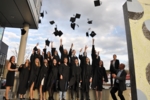Bild zeigt das gute Dutzend in Talare gekleidete Absovlventen vor dem Gebäude in Biberach, wie sie gerade ihre Hüte in die Höhe werfen und damit den Erwerb ihres akademischen Titels feiern. Erfreut schauen ihre Professoren zu.