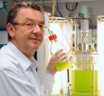 Zu sehen ist ein Mann mit Brille, der vor einem Bioreaktor in einem Labor sitzt und einen Kolben mit einer grünen flüssigkeit in der Hand hält.